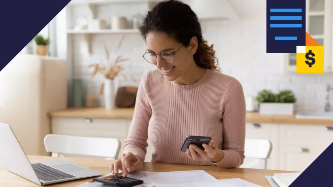 Mulher sorridente usando óculos, calculando finanças no lar com um laptop, calculadora e smartphone à mão. Ela está em um ambiente doméstico bem-iluminado e decorado de forma simples, demonstrando concentração e satisfação ao gerenciar seu orçamento.