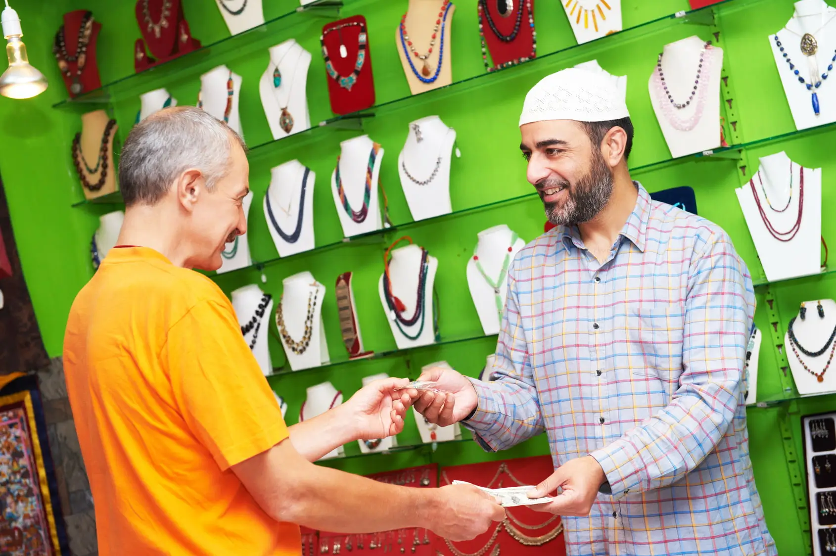 Dois homens em uma loja de joias. Um deles, vestindo camisa listrada e chapéu tradicional, sorri enquanto entrega joias ao cliente, que está de camisa laranja. Ao fundo, há colares exibidos em suportes brancos sobre uma parede verde.