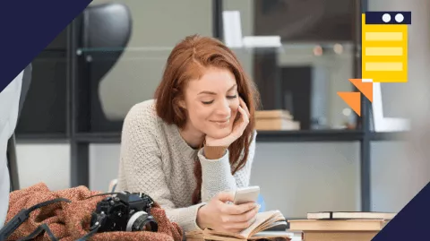 Uma mulher ruiva, sorridente e casualmente vestida, lê algo em seu smartphone enquanto se apoia com a mão no queixo, rodeada de livros e uma câmera fotográfica, indicando um ambiente criativo e descontraído.