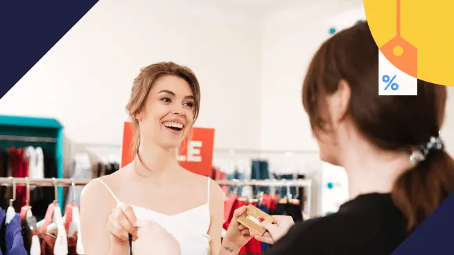 Duas mulheres em uma loja de roupas, uma segurando uma peça e sorrindo enquanto a outra oferece um cartão de crédito. Ao fundo, uma placa de "SALE". Ícone de etiqueta de preço com porcentagem no canto superior direito.