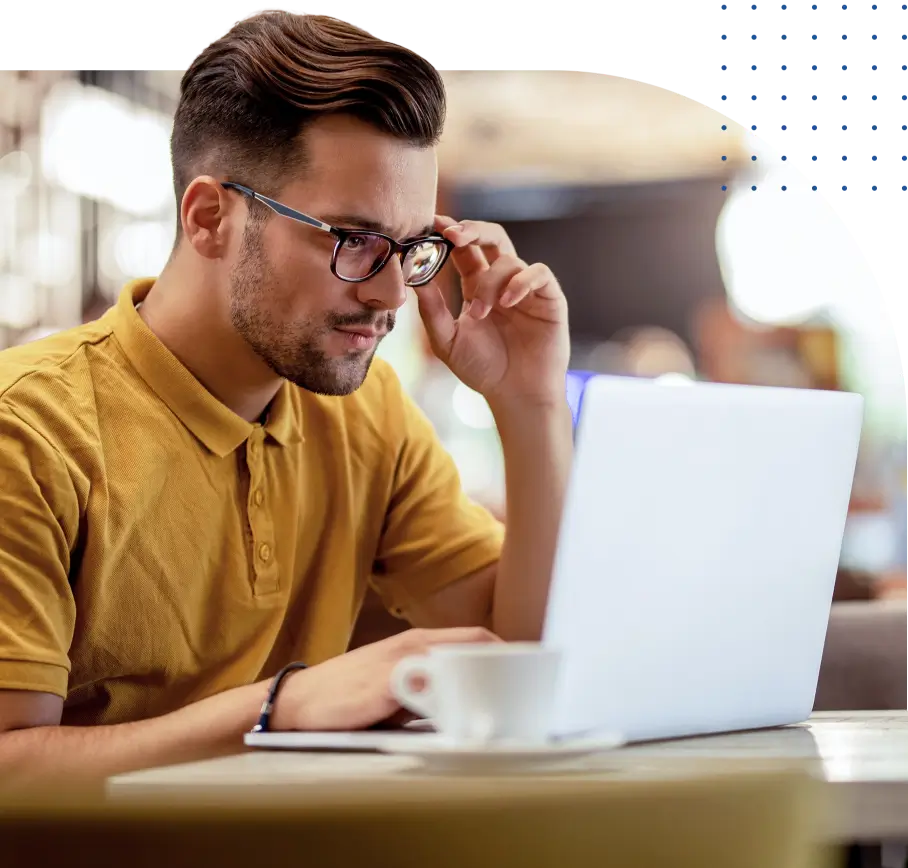 Um homem com barba, usando óculos e uma camisa polo amarela, está concentrado olhando para a tela de um laptop. Ele ajusta os óculos com uma mão. Na frente dele, há uma xícara de café branca.