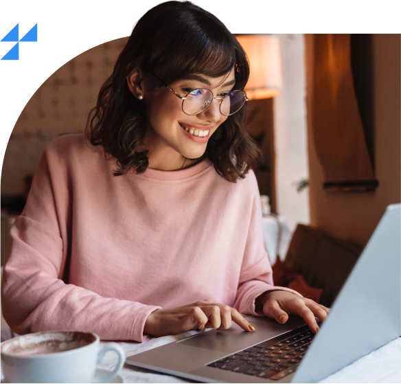 Mulher sorridente usando óculos e suéter rosa, digitando em um laptop. Ela está sentada em um ambiente aconchegante, com uma xícara de café ao lado. Ícone azul aparece no canto superior esquerdo.