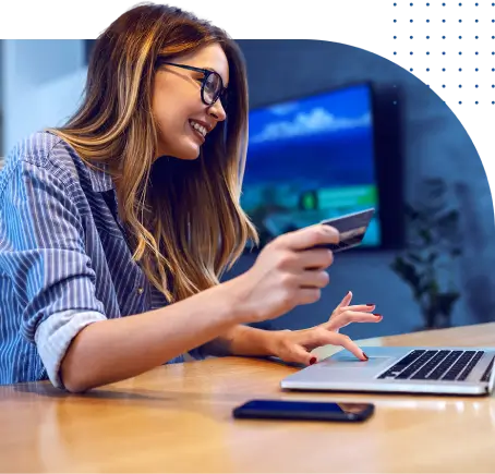 Mulher sorrindo, segurando um cartão de crédito enquanto usa um laptop. Um smartphone está sobre a mesa, e ao fundo, uma TV desfocada exibe uma paisagem. Pontos azuis no canto superior direito da imagem.